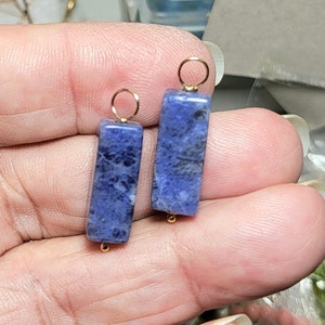 Puede incluir: Dos pendientes rectangulares de piedra sodalita azul con detalles dorados. Cada pendiente tiene un lazo dorado en la parte superior y una pequeña cuenta dorada en la parte inferior. Los pendientes se muestran sobre un fondo neutro.