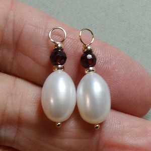 May include: A pair of jewelry charms featuring oval white pearls suspended below dark red faceted beads. Each charm includes a small gold-toned metal jump ring at the top and a tiny gold-toned metal bead at the base, creating a symmetrical design.