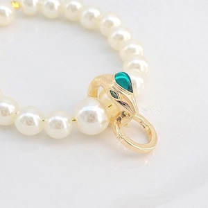 May include: A bracelet composed of round white faux pearls featuring a gold-toned metal clasp shaped like a snake head. The snake head is accented with a vibrant green enamel inlay on the top and small green enamel eyes.