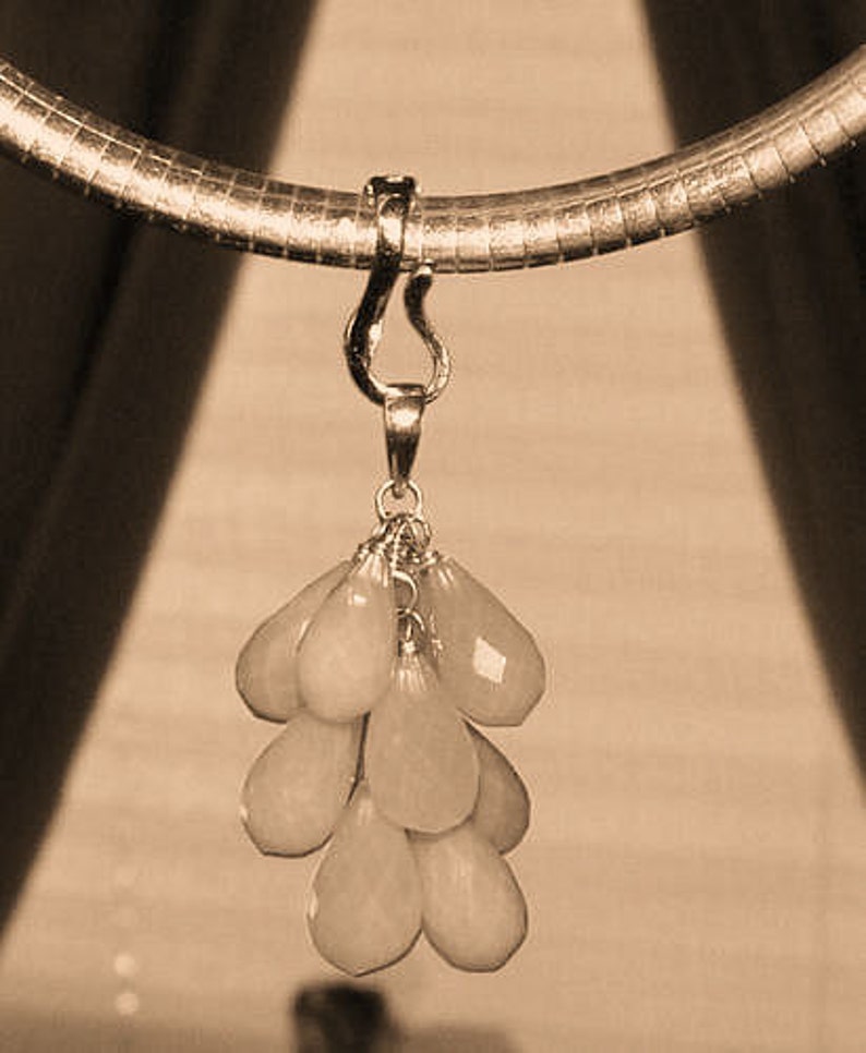 May include: A sepia-toned image of a necklace featuring a gold-colored chain and pendant. The pendant is a cluster of pale peach-colored, teardrop-shaped gemstones. The necklace is suspended against a neutral background.