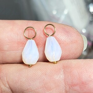 May include: Two small, faceted, pear-shaped white opalescent beads, each suspended from a thin gold-toned metal jump ring. The beads have a smooth, semi-translucent appearance with subtle blue and pink light reflections on their surfaces.