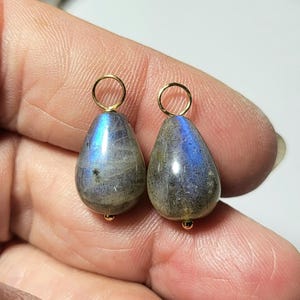 May include: Two teardrop-shaped labradorite gemstone pendants with gold-colored loops and small gold beads at the base. The stones have a mix of gray, blue, and iridescent colors. The pendants are held in a hand.