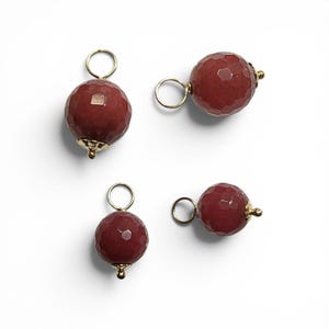 May include: Four spherical charms featuring faceted dark red stones. Each charm is topped with a gold-toned metal cap and a circular jump ring for attachment. The stones have a textured, multi-faceted surface that reflects light, creating a polished appearance.