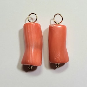 Puede incluir: Dos colgantes cilíndricos de coral color salmón, con una superficie lisa y pulida. Cada pieza presenta una textura ligeramente irregular con un sutil veteado blanco. Un pequeño anillo de metal dorado está unido a la parte superior.