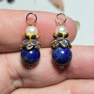 May include: Two matching pendants featuring a round, deep blue lapis lazuli stone at the base, topped with a gold-toned metal flower cap, a small white pearl, and a gold-toned jump ring at the top for attachment to a chain or earring hook.