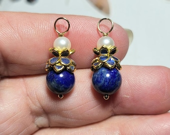 May include: Two matching pendants featuring a round, deep blue lapis lazuli stone at the base, topped with a gold-toned metal flower cap, a small white pearl, and a gold-toned jump ring at the top for attachment to a chain or earring hook.