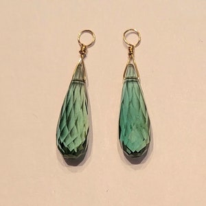May include: A pair of teardrop-shaped earrings with faceted, teal-colored glass pendants. The pendants are suspended from gold-colored wire loops. The earrings are displayed against a white background.
