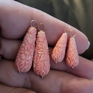 May include: Four coral-colored earrings with floral carvings. Two pairs of earrings are visible, each with a different size and design. The earrings have gold-colored hoops and small gold beads at the bottom.