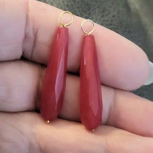 May include: Two faceted, teardrop-shaped, red-colored gemstone pendants with gold-colored loops. The pendants are held in a hand against a blurred background. The gemstones have a polished, shiny appearance.