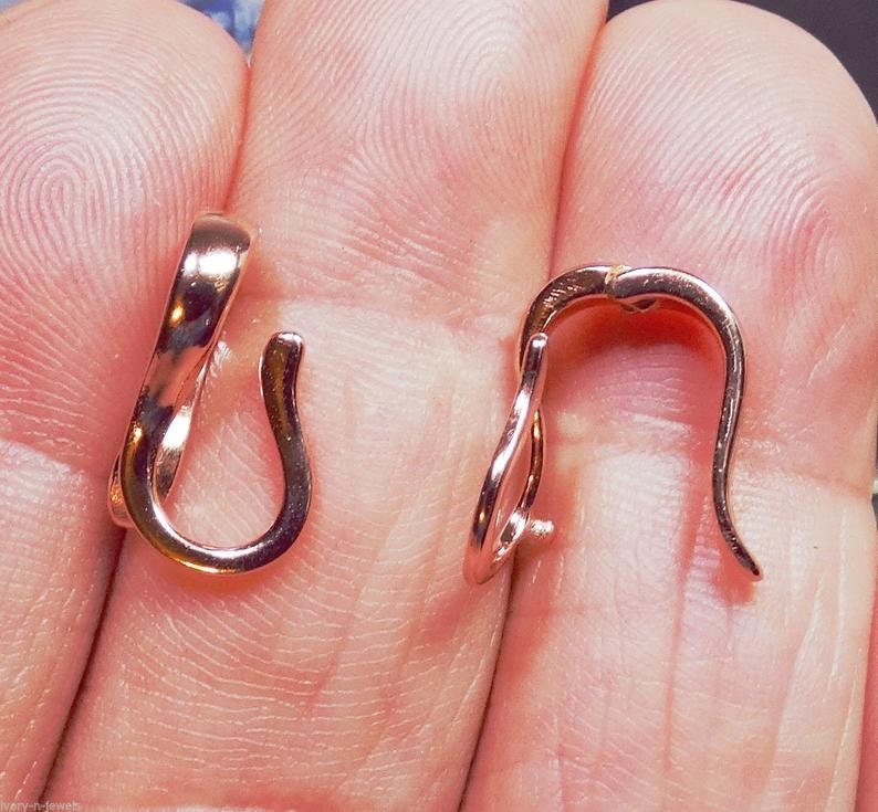 May include: Two rose gold-colored earring hooks are displayed. The hooks have a curved design with a small loop at the bottom and a curved top. The hooks are shown against a skin-toned background.