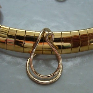 Könnte beinhalten: Eine goldfarbene Halskette mit einem tropfenförmigen Anhänger. Die Halskette hat ein geripptes Design, und der Anhänger ist eine Doppelschleife. Der Schmuck wird auf einer hellgrauen Oberfläche präsentiert.