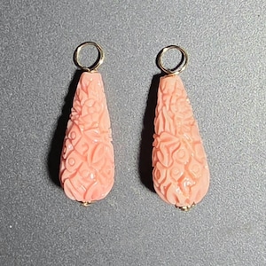 May include: A pair of teardrop-shaped pendants in a soft peach color, each featuring intricate floral carvings across the surface. Each pendant is topped with a small, circular gold-toned metal jump ring for attachment to a chain or earring hook.