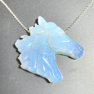 May include: A pendant shaped like a horse head carved from a translucent, pale blue opal-like stone. The pendant is suspended from a thin, silver-colored metal chain. The carving features detailed lines representing the horse's mane and facial features.