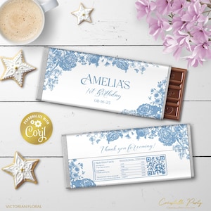 May include: A white chocolate bar wrapper with a blue floral design. The wrapper has the text "Amelia's 1st Birthday 08-16-25" and "Thank you for coming!" The wrapper also includes a QR code.