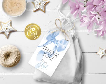 Può includere: Un sacchetto regalo bianco con un'etichetta a fiocco blu con la scritta "THANK YOU!" e "Love, Elyah". L'immagine include una tazza di caffè, biscotti a forma di stella, un gomitolo di spago e fiori rosa, il tutto su una superficie di legno bianco.