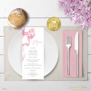 Może przedstawiać: Eleganckie nakrycie stołu z białym talerzem, różową serwetką i sztućcami. Karta menu z różowym wzorem kokardy i napisem "Menu" znajduje się na talerzu. Bułka i różowe kwiaty dopełniają aranżację.