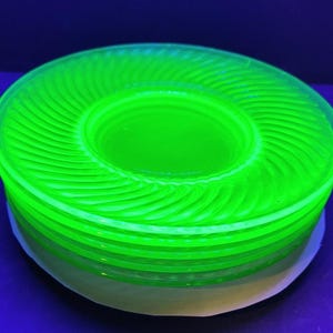 May include: A stack of bright neon green glass plates. Each plate has a spiral design radiating from the center. The plates are stacked on a white surface, with a dark blue background.