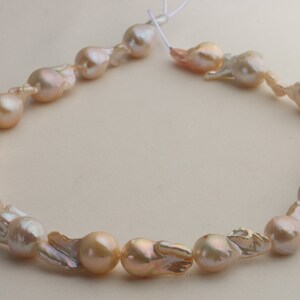 May include: A strand of irregularly shaped, iridescent freshwater pearls. The pearls display a range of colors, including cream, peach, and hints of pink and brown. The pearls are strung together with a thin, white string, creating a necklace.