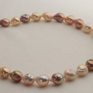 May include: A strand of baroque pearls in shades of pink, cream, and white. The pearls are strung on a white cord and are arranged in a circular pattern.