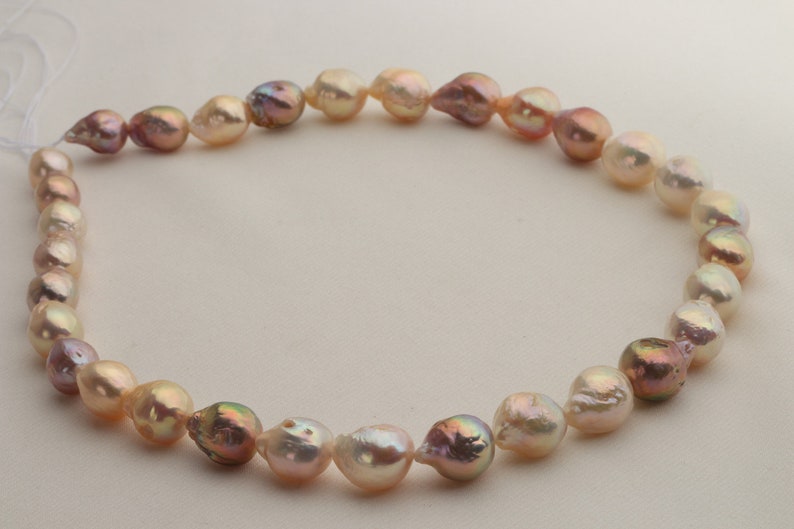 May include: A strand of baroque pearls in shades of pink, cream, and white. The pearls are strung on a white cord and are arranged in a circular pattern.