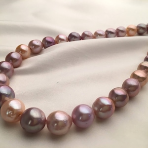 Puede incluir: Una hebra de perlas con un degradado de colores rosa, morado y blanco. Las perlas están enhebradas en un hilo fino y están dispuestas en un patrón circular.