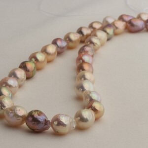 May include: A strand of baroque pearls in shades of pink, peach, and cream. The pearls are strung on a white thread and are arranged in a loose, flowing pattern.