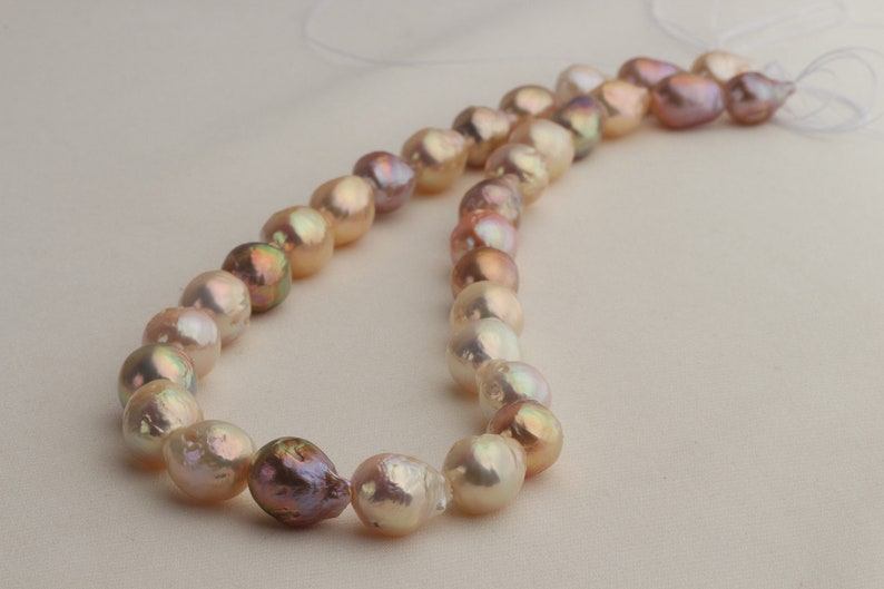 May include: A strand of baroque pearls in shades of pink, peach, and cream. The pearls are strung on a white thread and are arranged in a loose, flowing pattern.