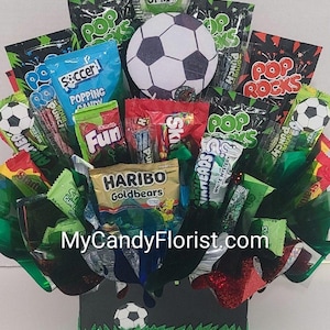 May include: A candy bouquet featuring various snack packages including Haribo Goldbears, Pop Rocks, and Skittles, arranged with a central soccer ball graphic and a black base with a soccer ball and green grass design, labeled MyCandyFlorist.com.