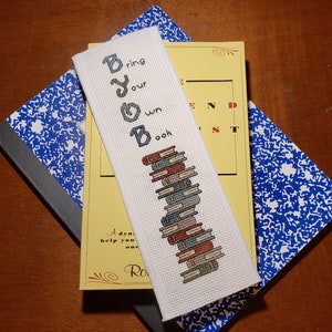 May include: White bookmark with cross-stitch embroidery that reads "Bring Your Own Book" and a stack of colorful books.