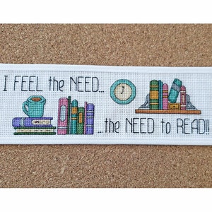 Op de afbeelding: Witte boekenlegger met borduurwerk in kruissteek, met daarop een kopje koffie, een stapel boeken, een boekenkast met boeken en een klok. De tekst luidt "I FEEL the NEED... the NEED to READ!"