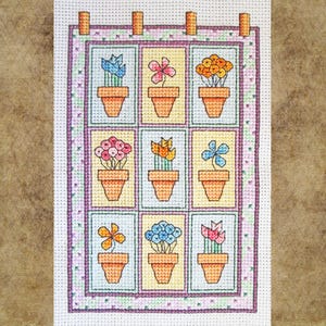 May include: A cross-stitch wall hanging with nine squares, each displaying a potted flower in various colours. The flowers include blue, pink, and orange blooms. The piece is framed with a purple border and has wooden dowels at the top for hanging.