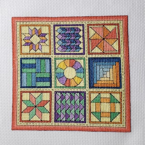 May include: A colorful cross-stitch embroidery of nine different quilt block patterns. The blocks are arranged in a 3x3 grid and are framed with a red border.
