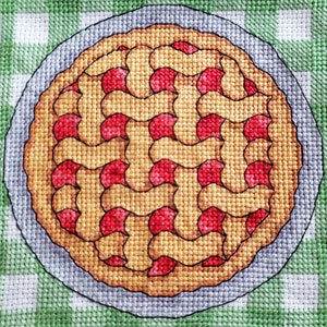 May include: A close-up of a cross-stitch design of a cherry pie with a lattice crust. The pie is on a green and white checkered background.