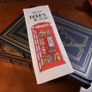 May include: A white bookmark with a red telephone booth design. The booth is filled with books and the text "Your Books Are Calling!" is printed above the booth.