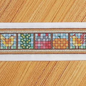 May include: A cross-stitch bookmark with a colorful patchwork design featuring a bird, a house, a pumpkin, and a flower. The bookmark is framed in brown and has a white background.