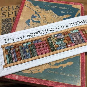 Könnte beinhalten: Ein weißes Kreuzstich-Lesezeichen mit einem Bücherregal-Design und dem Text "It's not HOARDING if it's BOOKS!". Das Lesezeichen liegt auf mehreren alten Büchern mit abgenutzten Einbänden.