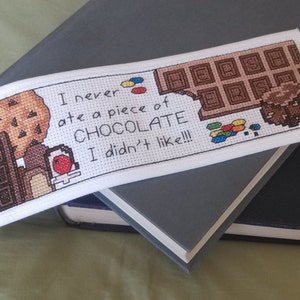 Op de afbeelding: Witte bladwijzer met borduurwerk in kruissteek, met daarop een chocolade reep, een koekje en een klein persoon. De tekst luidt "Ik heb nog nooit een stuk CHOCOLADE gegeten dat ik niet lekker vond!!!"