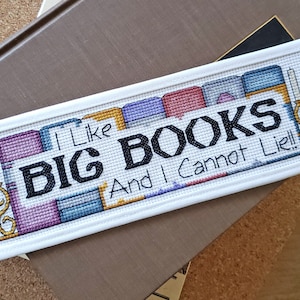 May include: A white bookmark with a cross-stitch design featuring a stack of colorful books and the text "I Like Big Books And I Cannot Lie!"