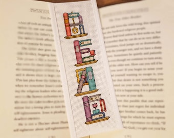 May include: A white bookmark with a cross-stitch design of a bookshelf with books and other decorative items. The word "READ" is spelled out using the books on the shelf.