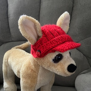 May include: A plush dog wearing a red knitted hat with a black button. The hat has a brim and is pulled down over the dog's ears.