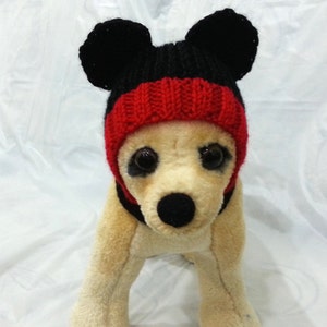 May include: A stuffed dog, in shades of tan, wearing a knitted hat in black, red, and black, with large, round ears. The hat has a red band around the forehead. The dog has large, dark eyes and a black nose.