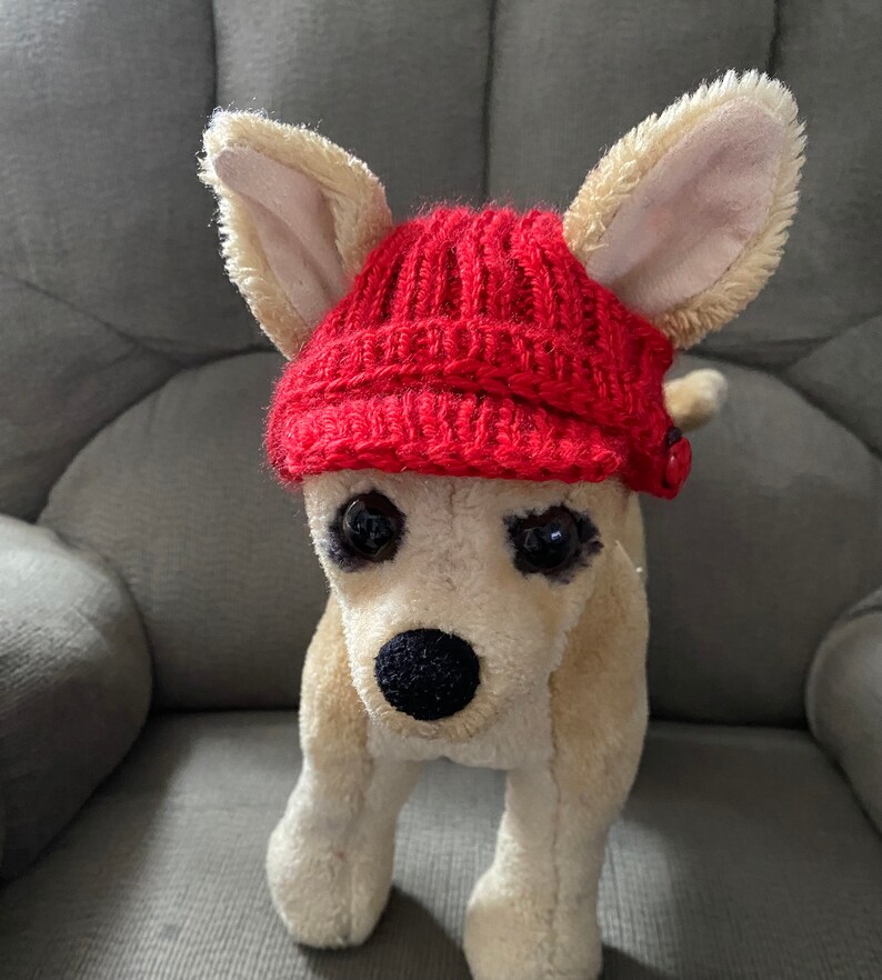 May include: A plush toy dog wearing a red knitted hat with a button on the side.