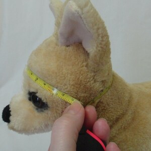 May include: A beige plush toy dog with black eyes and a black nose. A yellow measuring tape is wrapped around the dog's neck, measuring 12 inches.