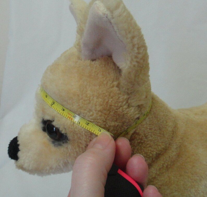 May include: A beige plush toy dog with black eyes and a black nose. A yellow measuring tape is wrapped around the dog's neck, measuring 12 inches.