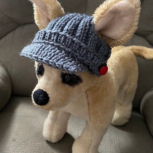 May include: A plush toy dog wearing a gray knitted hat with a brim. The hat has a red button on the side.