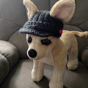 May include: A plush toy dog wearing a black knitted hat with a red button. The dog is sitting on a gray couch.