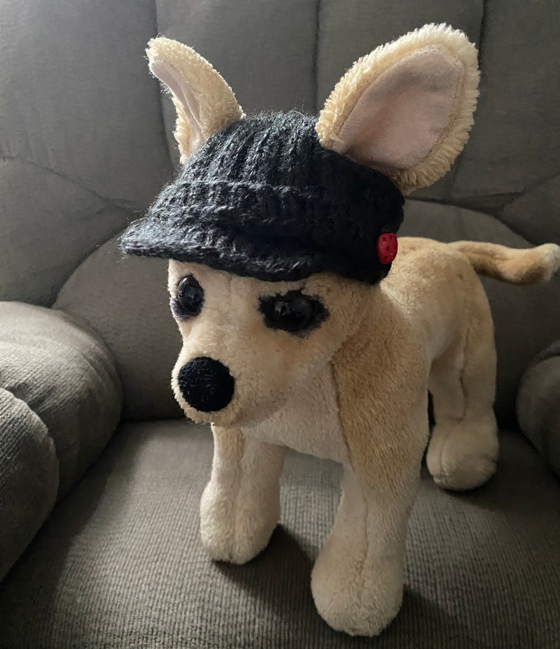 May include: A plush toy dog wearing a black knitted hat with a red button. The dog is sitting on a gray couch.
