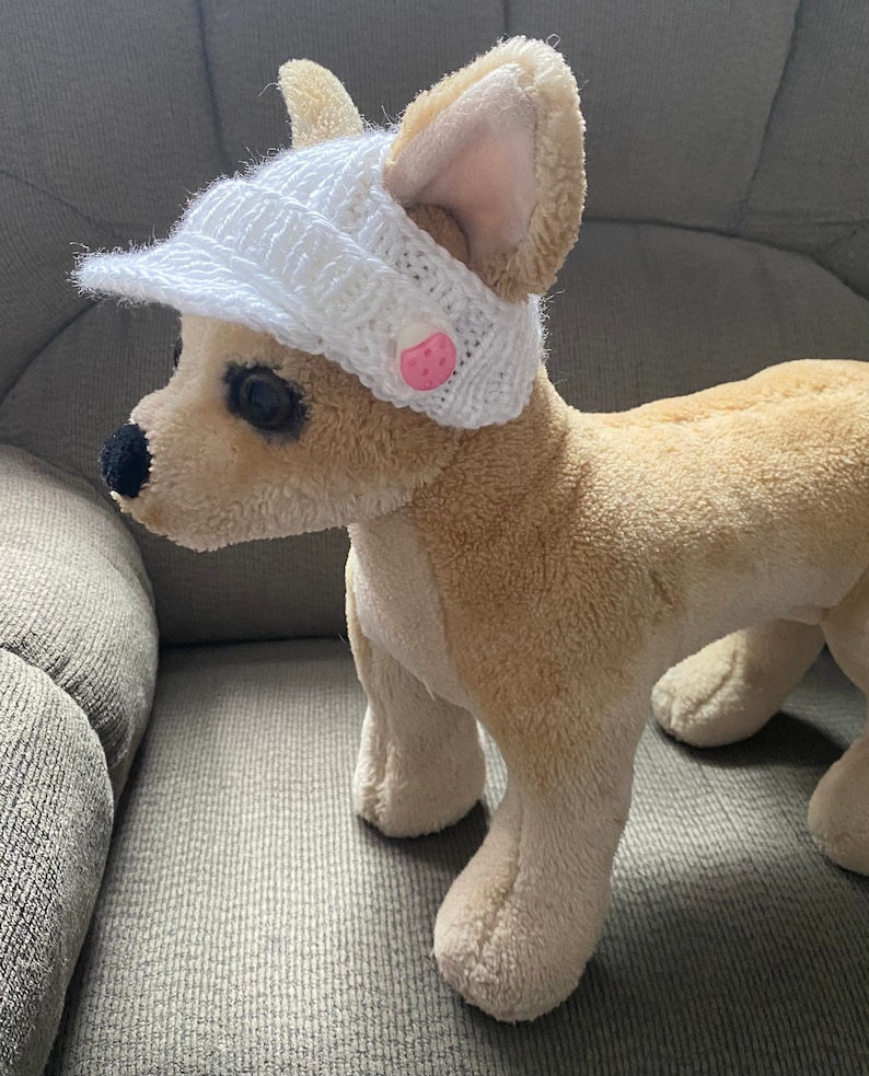 May include: A small, white crocheted hat with a pink button on the side. The hat is worn by a small, light brown stuffed dog.
