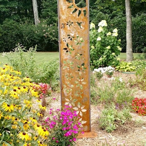 May include: A rusty metal garden sculpture with cutouts of flowers and a sun. The sculpture is standing in a garden with pink, yellow, and white flowers.