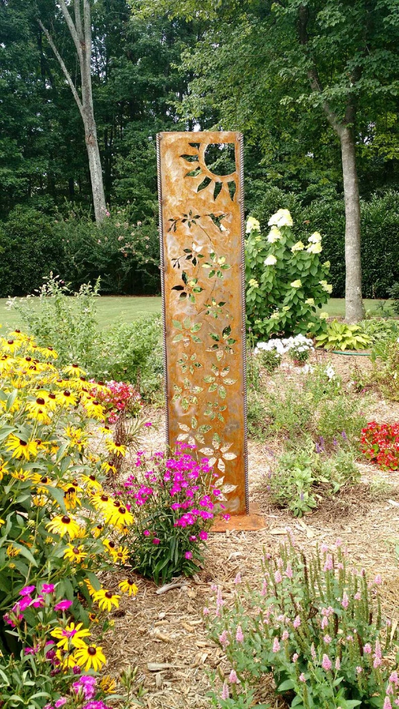 May include: A rusty metal garden sculpture with cutouts of flowers and a sun. The sculpture is standing in a garden with pink, yellow, and white flowers.